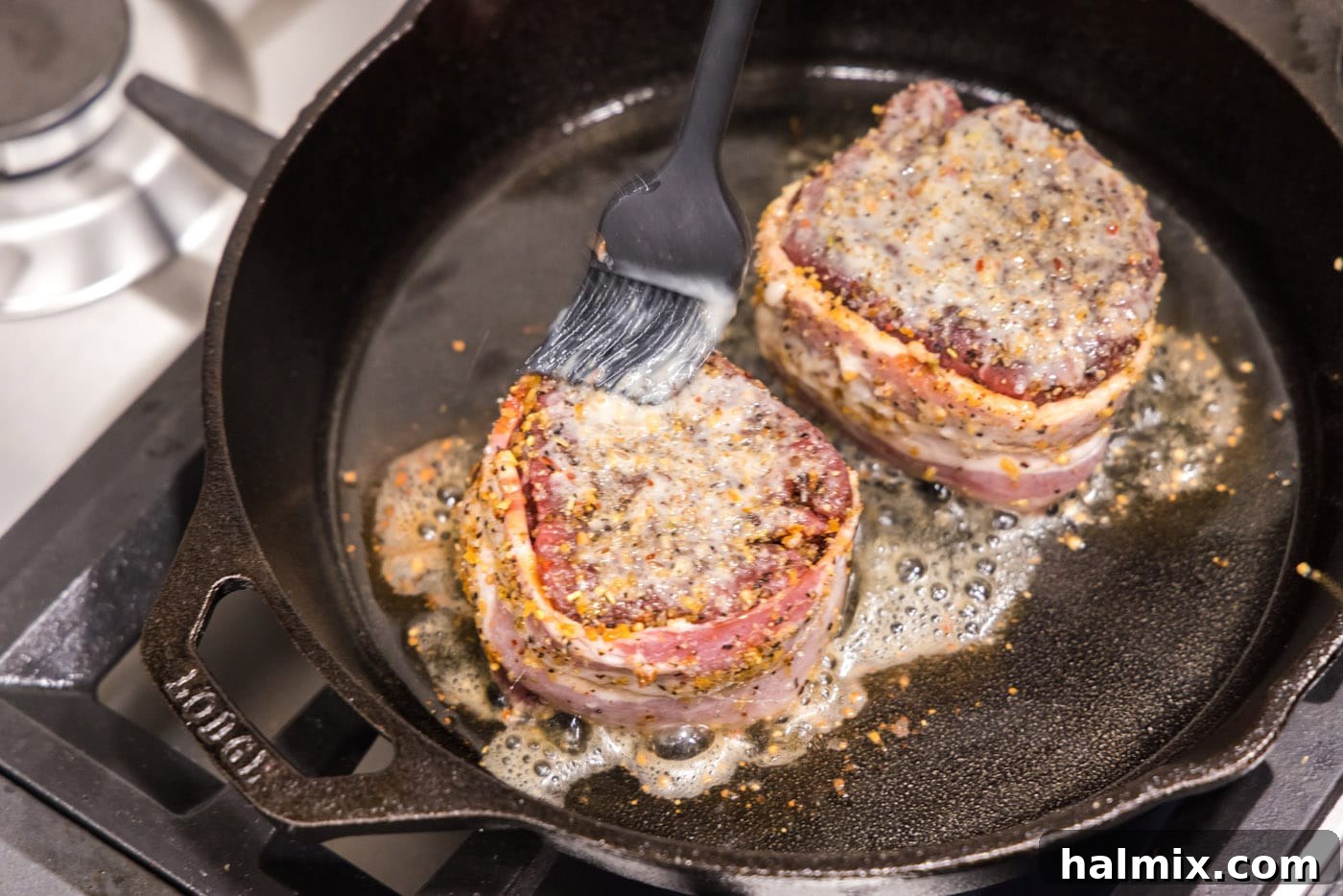 A close-up shot of a hand brushing butter onto a searing bacon-wrapped filet mignon in a skillet.
