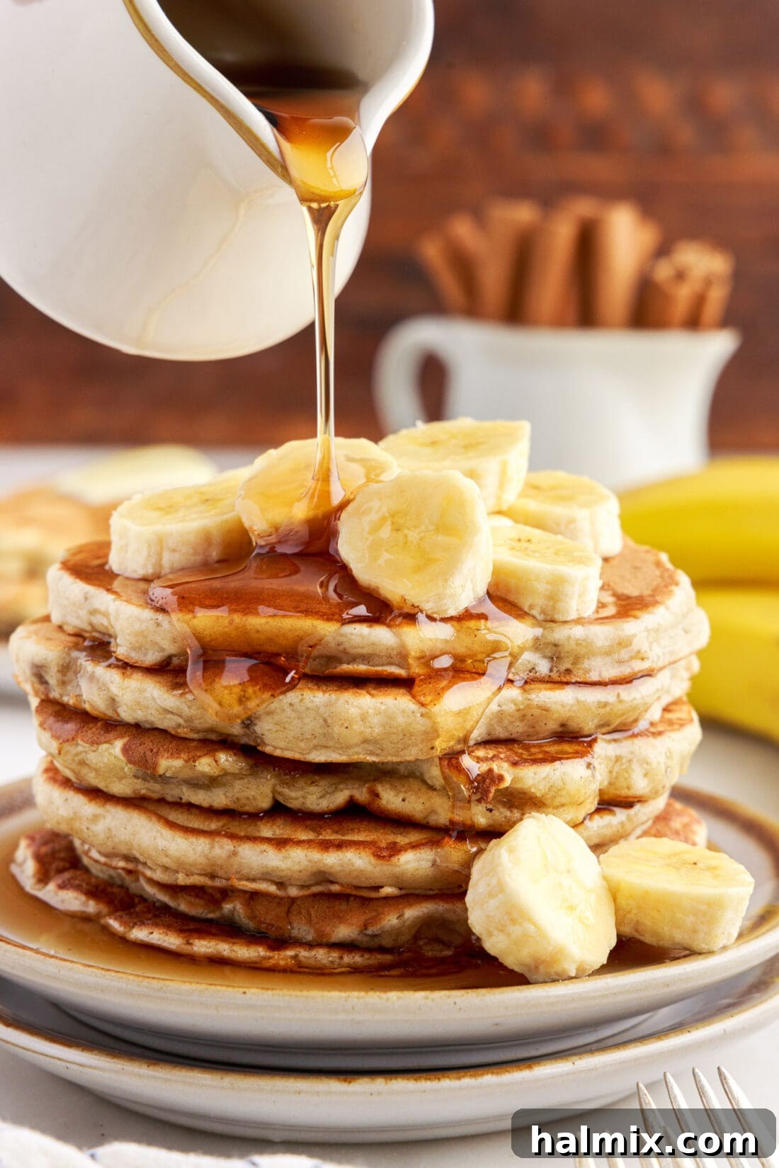 A tall stack of golden-brown fluffy banana pancakes, generously drizzled with rich maple syrup cascading down the sides