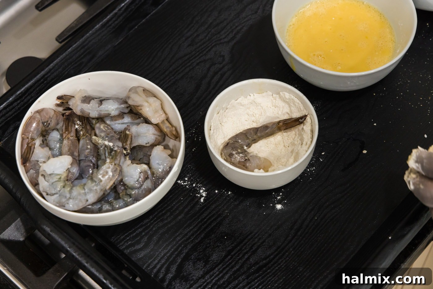 A hand dipping a plump shrimp into a bowl of flour