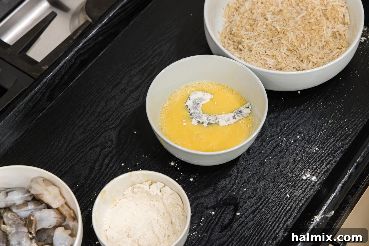 A hand dipping a floured shrimp into the whisked egg wash