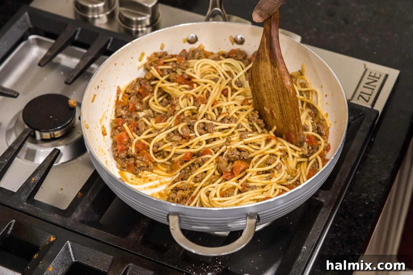 Mixing cooked spaghetti noodles into the flavorful taco seasoned ground beef, tomatoes, and onion sauce in a large skillet.