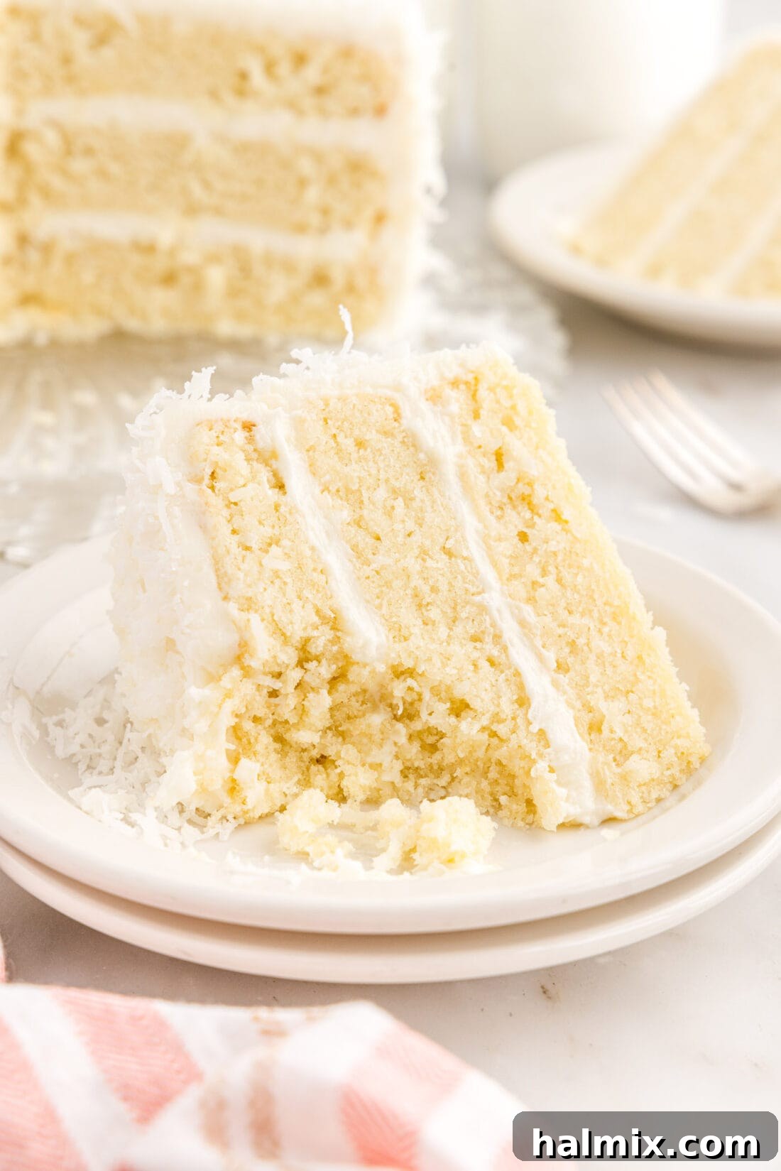 A delectable slice of moist coconut cake, beautifully frosted with cream cheese and topped with shredded coconut, resting on a plate with a small bite taken, highlighting its inviting texture.