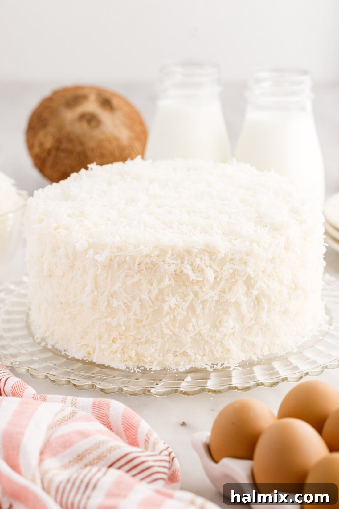 A pristine, three-layer homemade coconut cake, generously frosted with cream cheese and completely covered in shredded coconut, sits elegantly on a decorative cake stand, ready to be served.