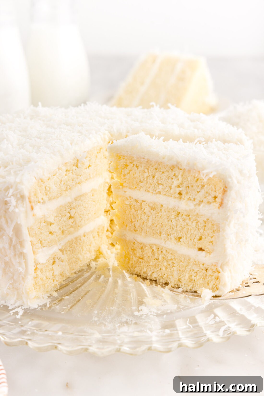 A stunning, generously frosted coconut cake on a white cake plate, with several slices already removed, revealing the moist layers and inviting texture within.