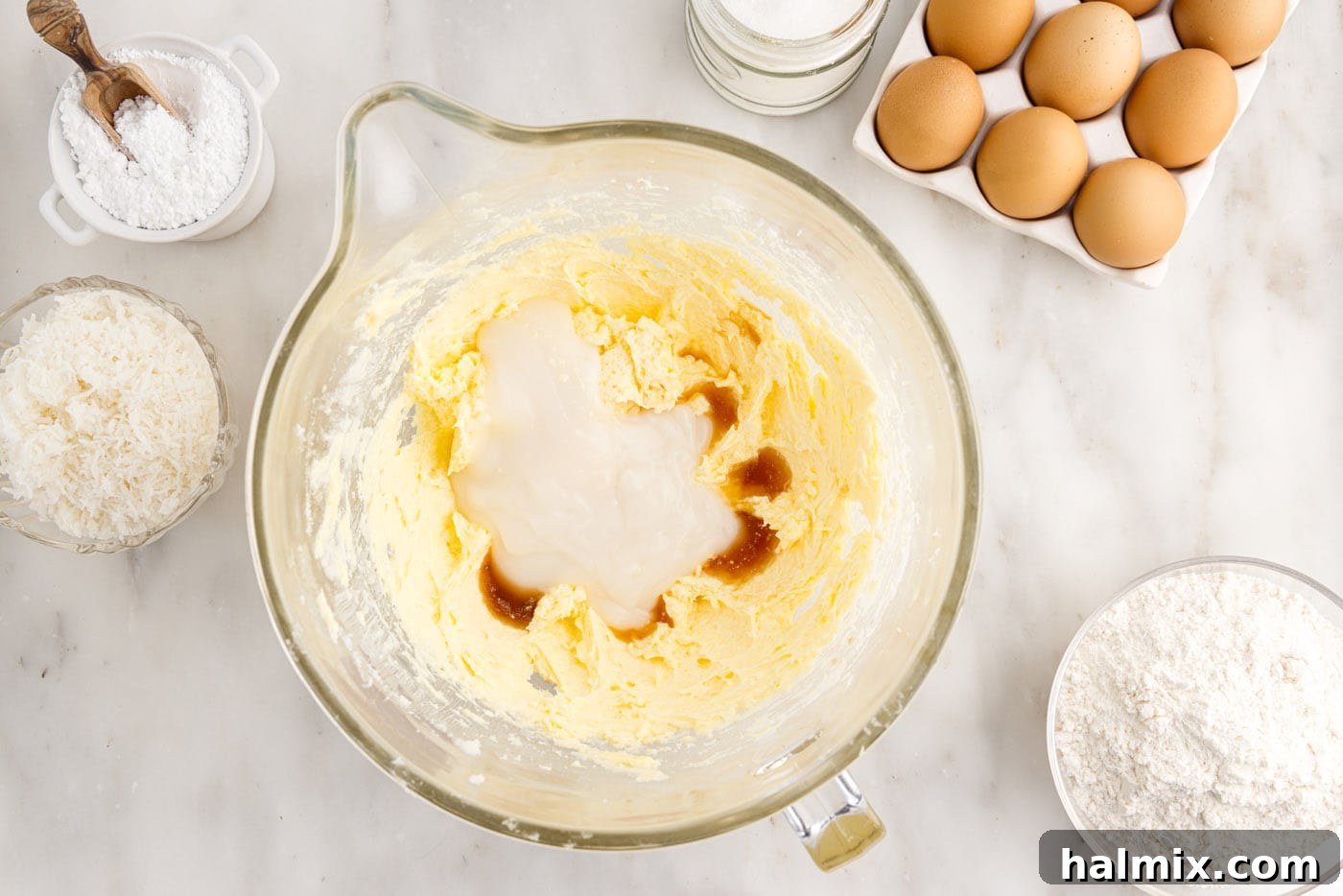 Coconut extract, vanilla extract, and cream of coconut being added to the cake batter in a mixing bowl, ready to be beaten for rich flavor.