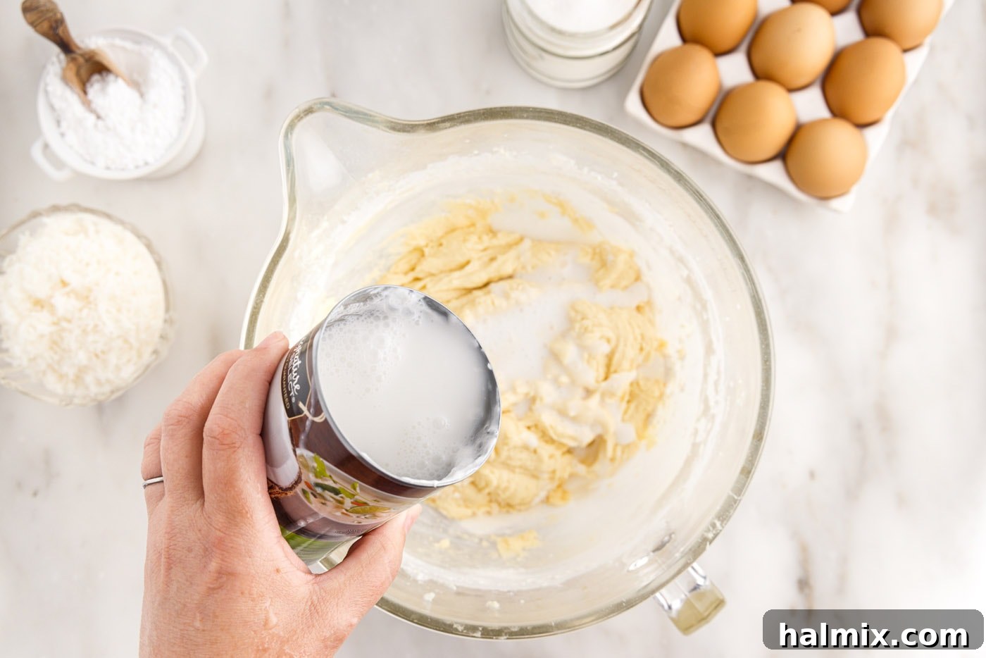 Unsweetened coconut milk being poured into the cake batter, alternating with dry ingredients for a perfectly mixed consistency.