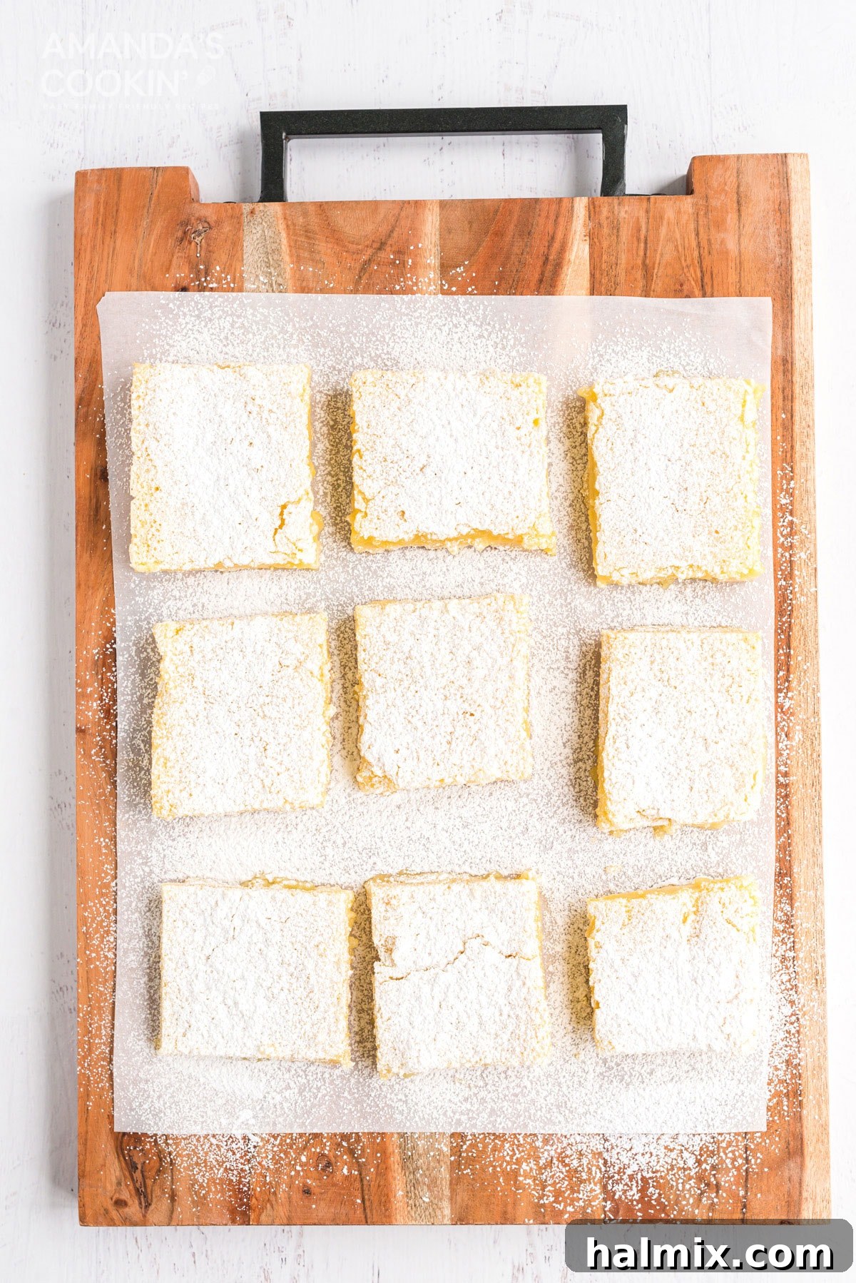 An inviting overhead view of freshly baked lemon bars neatly arranged on a wooden cutting board, showcasing their golden crusts and glossy, pale yellow tops dusted with powdered sugar. Ready to be sliced and served.