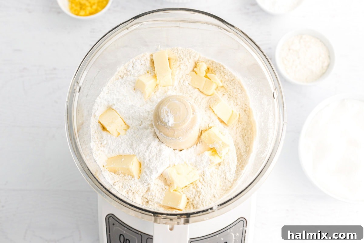 A food processor bowl filled with flour, powdered sugar, and cubed butter, ready to be processed into the shortbread crust mixture.