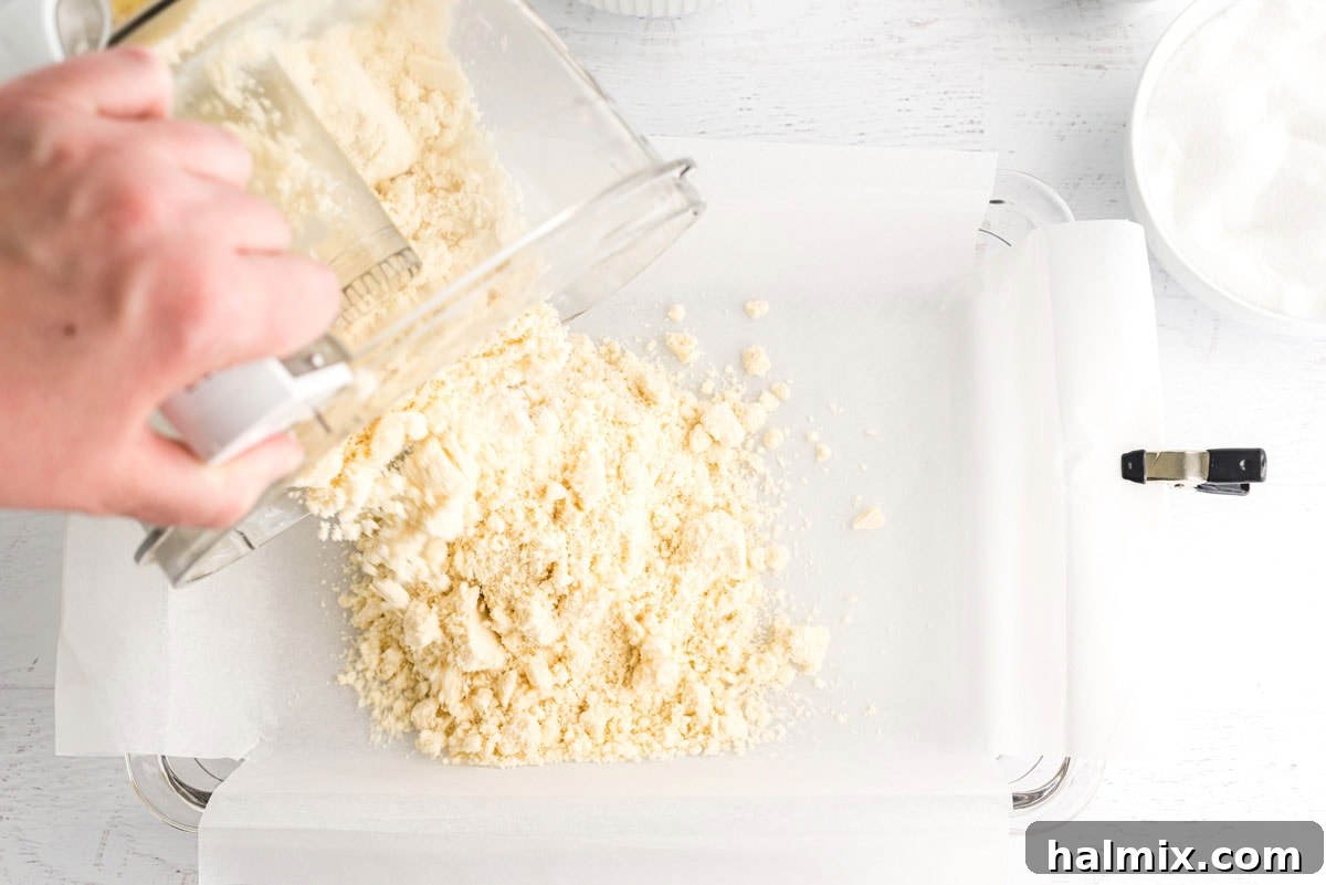 A close-up shot of the shortbread crumb mixture being poured into a parchment-lined 13x9 inch baking pan, ready to be pressed into an even crust.