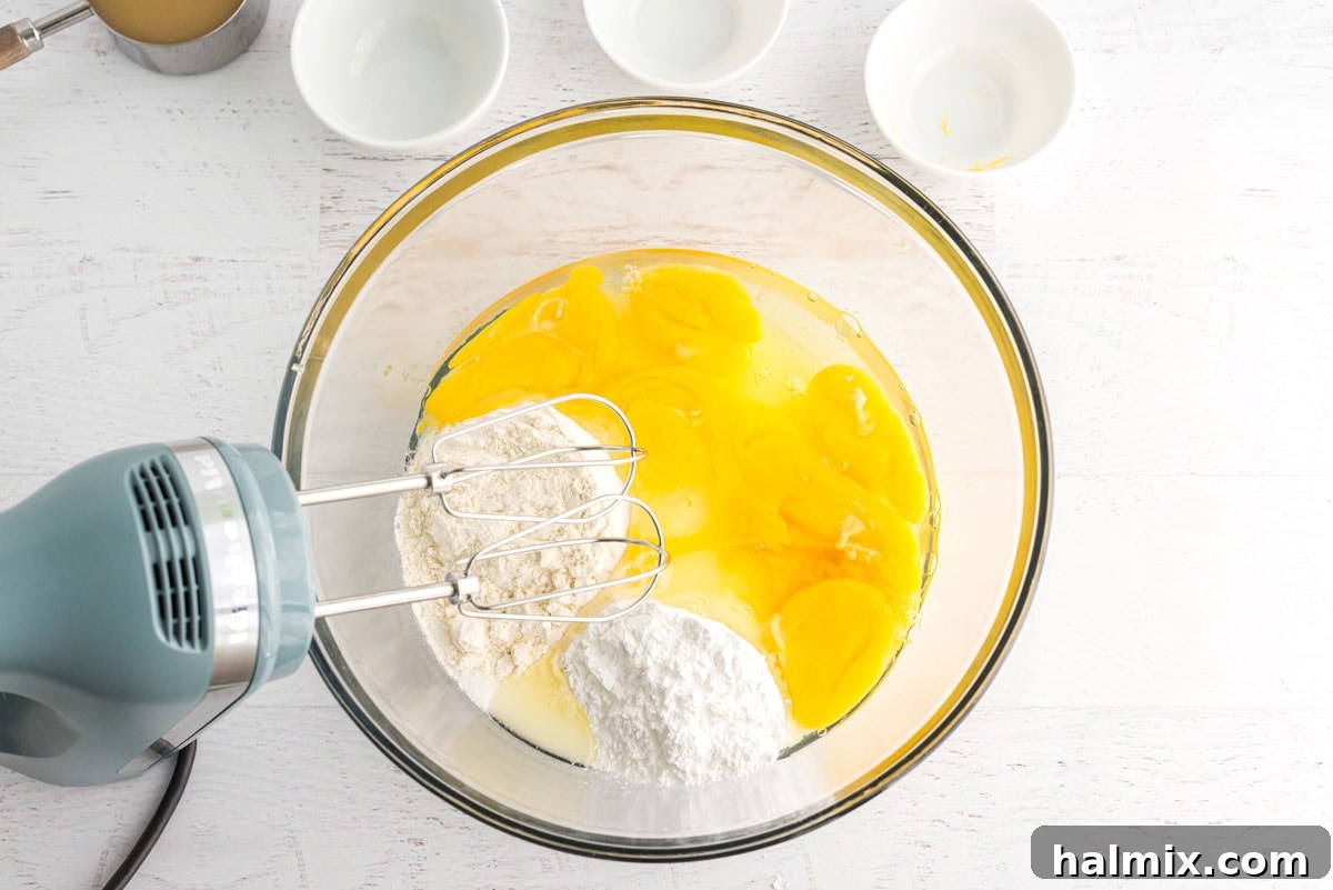 A mixing bowl with eggs, granulated sugar, flour, lemon juice, and lemon zest being beaten together by an electric mixer, creating a smooth, pale yellow mixture for the lemon bar filling.