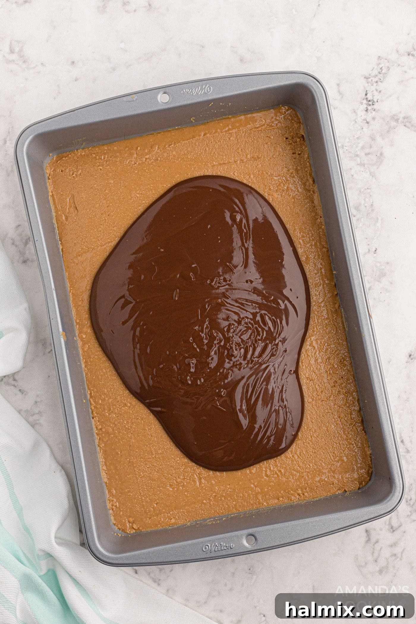 Melted chocolate and peanut butter spread over the golden peanut butter bar base in a baking pan, ready for chilling.