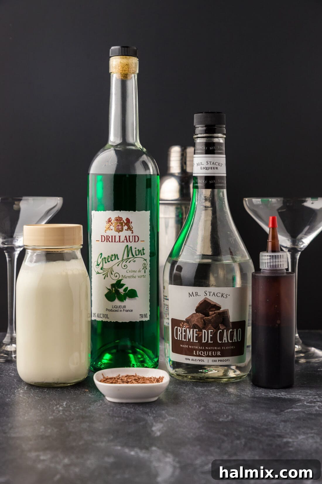 A collection of ingredients laid out for a Grasshopper drink: bottles of creme de cacao, creme de menthe, and a carton of heavy cream.