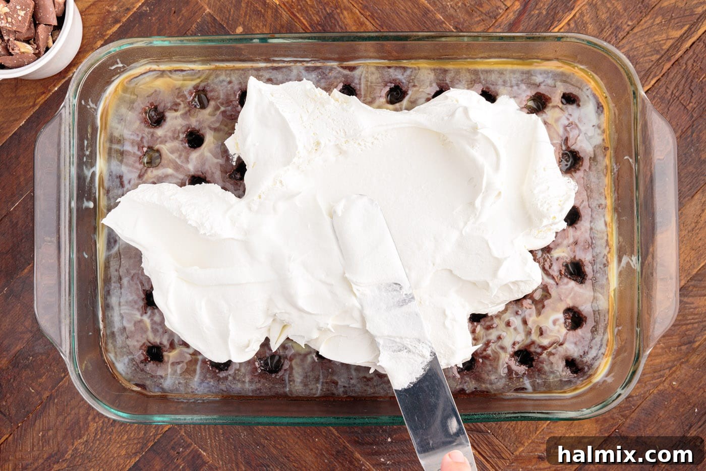 Fluffy Cool Whip topping being spread smoothly and evenly over the caramel-soaked chocolate poke cake, creating a pristine white layer.