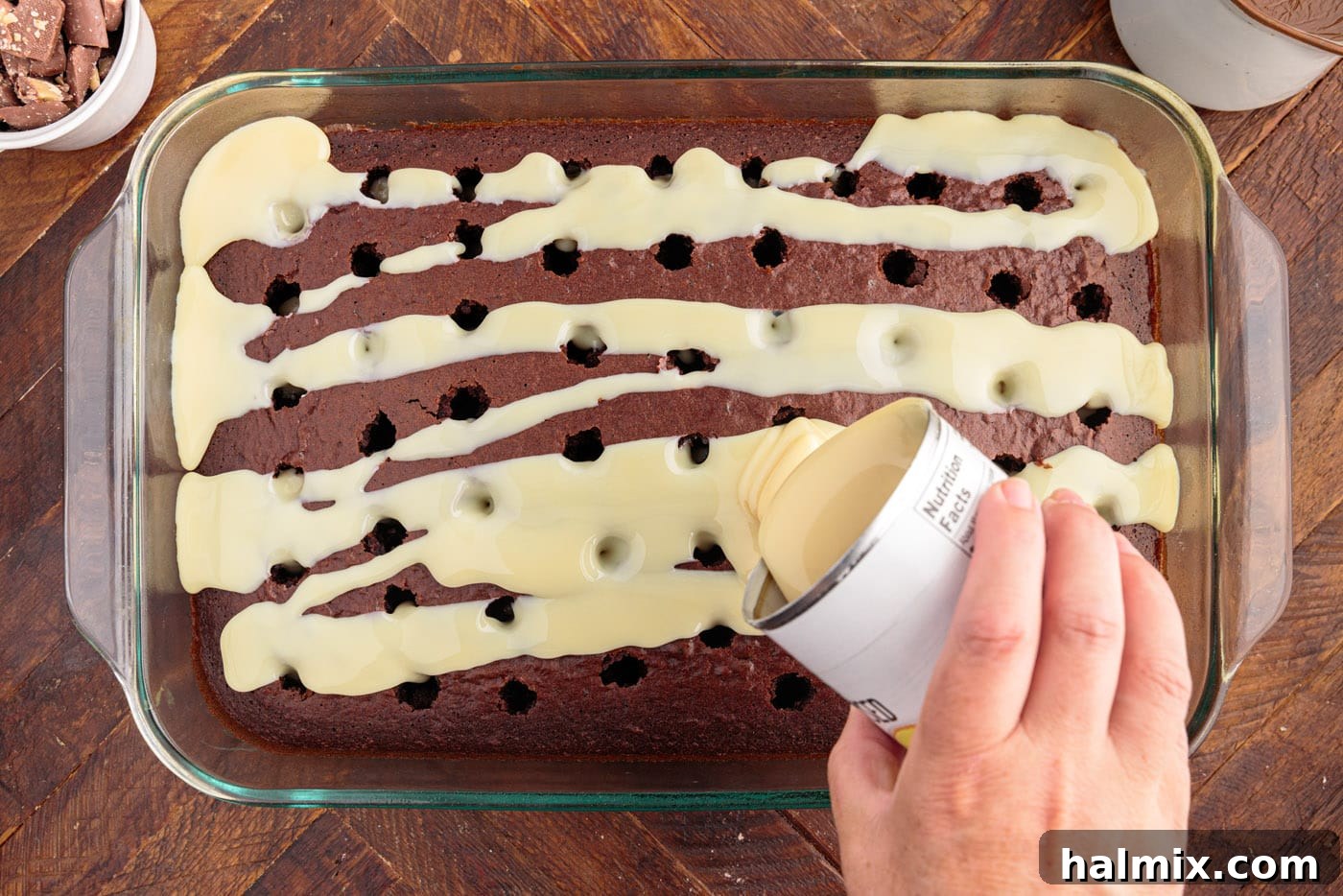 Sweetened condensed milk being generously poured over the poked holes of the chocolate cake, ready to soak in.