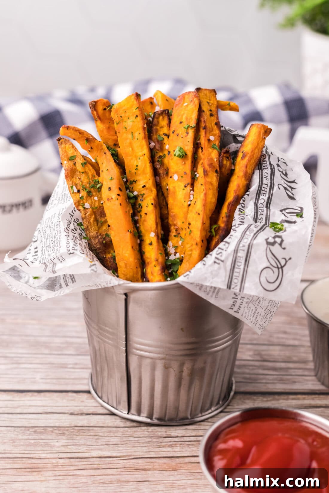 A serving of Air Fryer Sweet Potato Fries in a metal fry cup lined with parchment paper, accompanied by a small bowl of dipping sauce.
