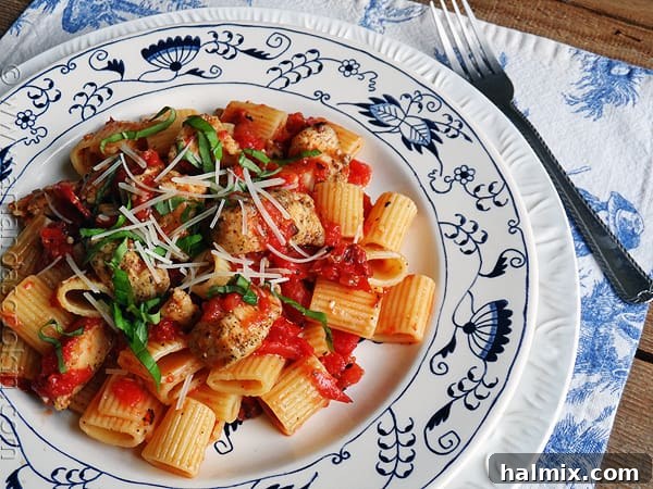 An overhead of a plate of spiced chicken and mezzi rigatoni topped with grated Parmigiano-Reggiano cheese and fresh basil.