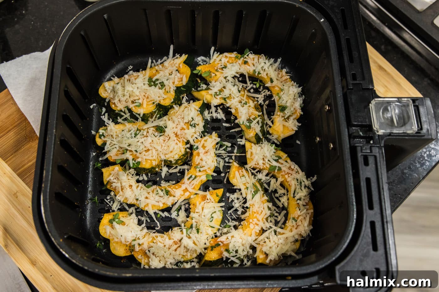 Shredded parmesan cheese generously added to sliced acorn squash in an air fryer basket, ready for cooking.