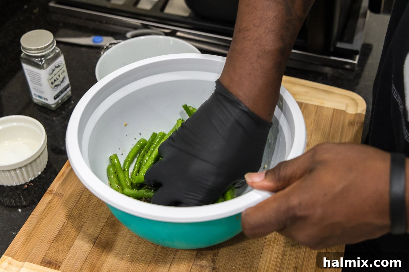 A gloved hand tossing fresh green beans with the olive oil and seasoning mixture in a large bowl, ensuring every bean is coated.