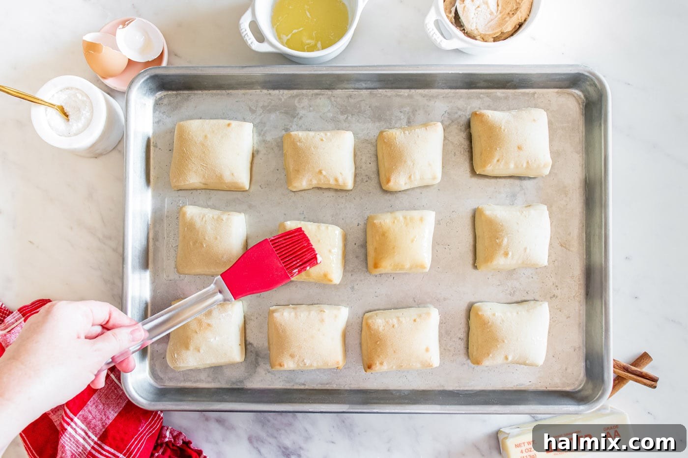 A basting brush applying melted butter to warm Texas Roadhouse rolls on a baking sheet.