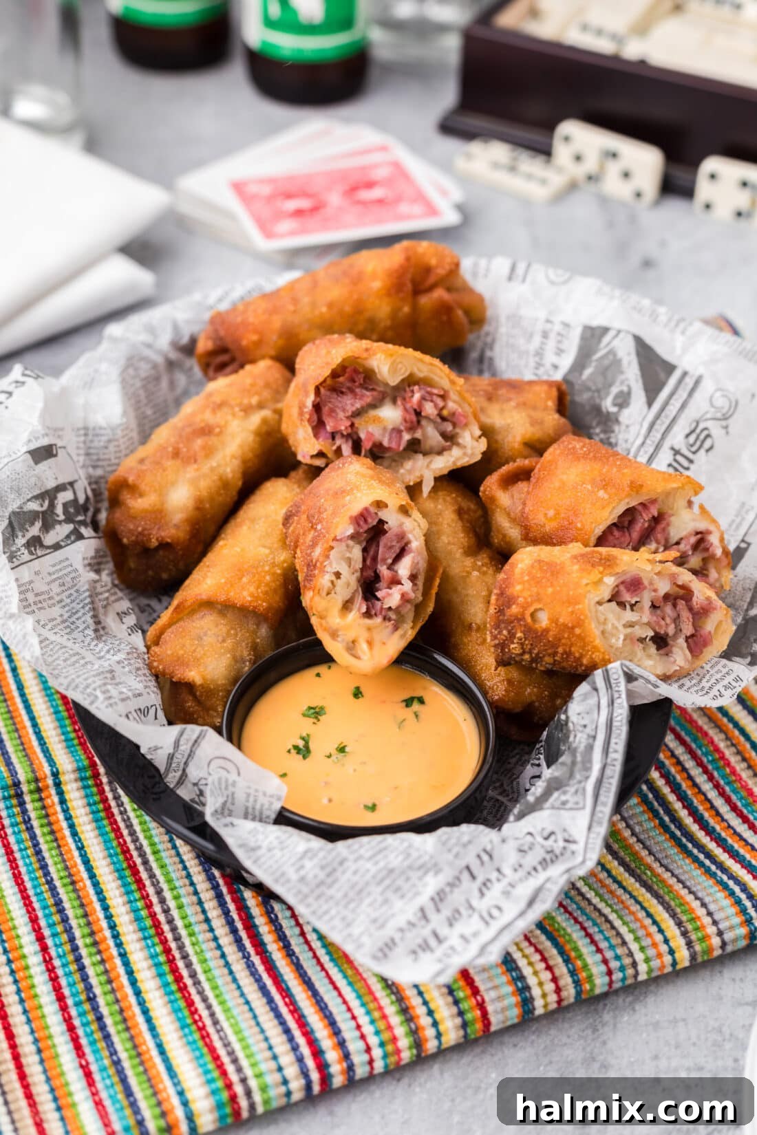 Crispy Reuben Egg Rolls 12 Reuben Egg Rolls cut in half, revealing the layers of corned beef, Swiss cheese, and sauerkraut, resting in a woven basket with a side of dipping sauce.