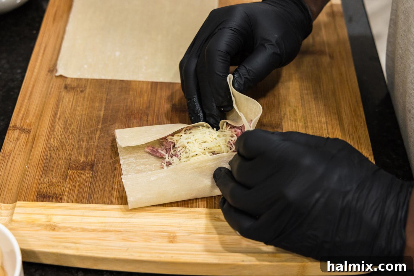 Hands carefully folding the bottom corner of an egg roll wrapper up and over the corned beef and Swiss cheese filling, demonstrating the initial tuck.