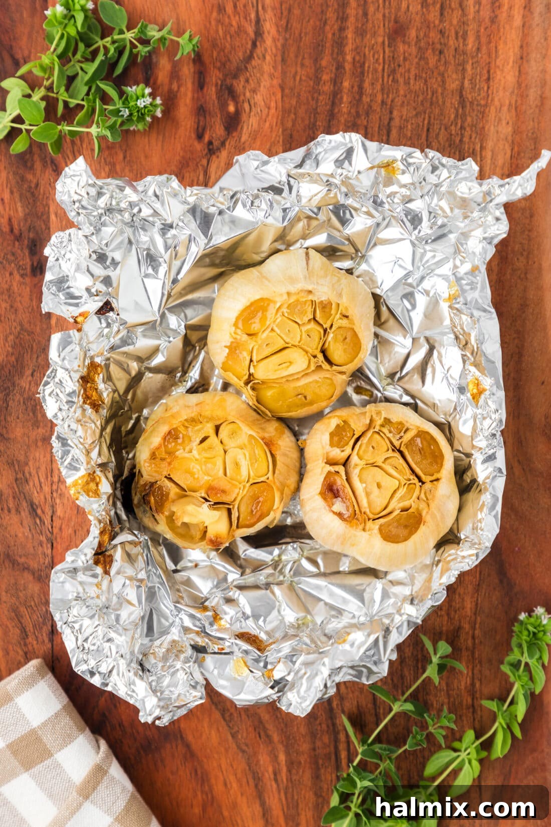 Three bulbs of perfectly roasted garlic, nestled in their foil wrapping, resting on a rustic wooden board, ready to be enjoyed.