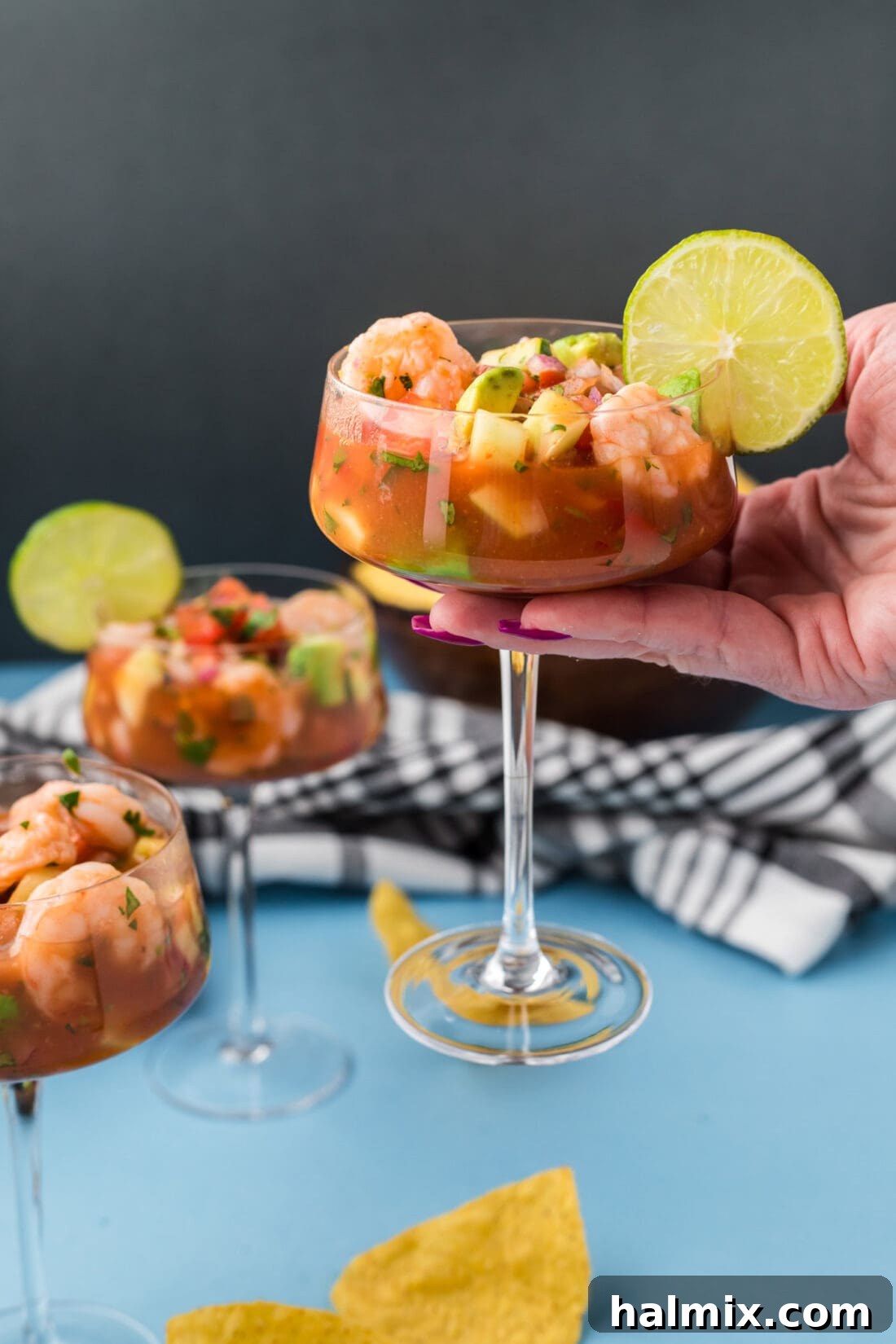 A hand holding a martini glass filled with a colorful Mexican Shrimp Cocktail, garnished with a lime wedge, against a blurred background