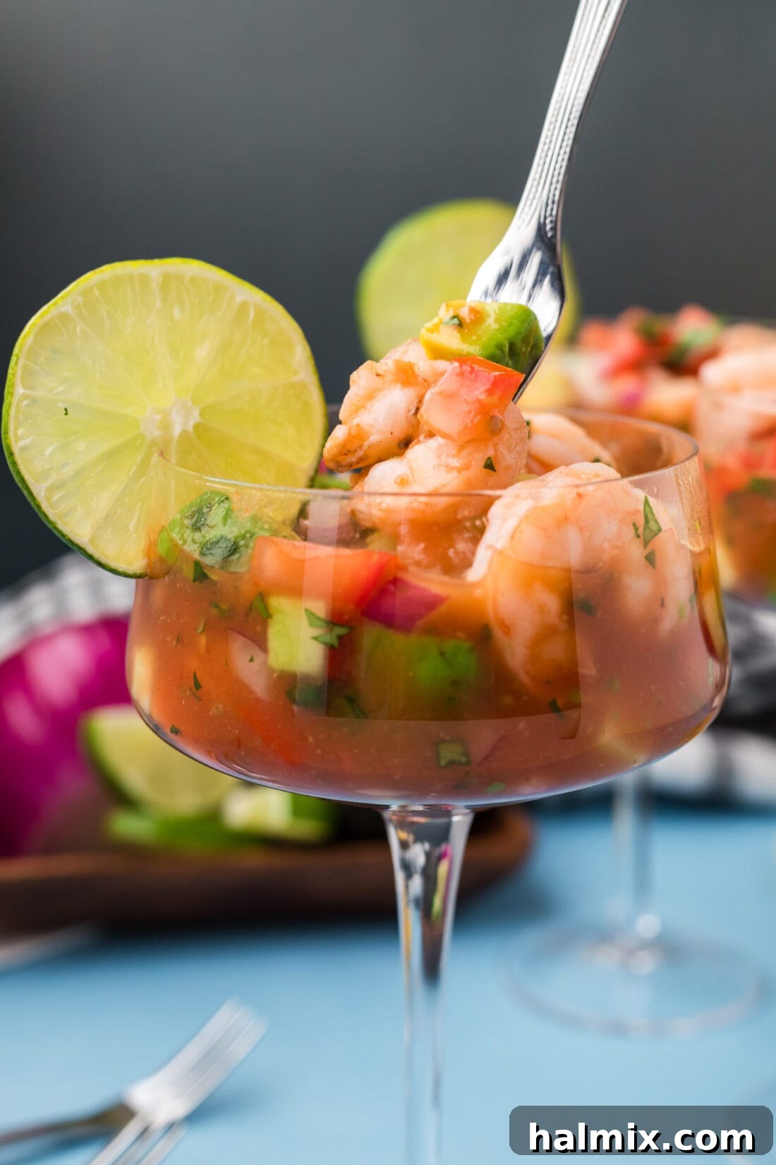 Close-up photo of a single Mexican Shrimp Cocktail served in a glass, with a fork dipped in, showcasing the fresh shrimp, diced vegetables, and rich tomato broth