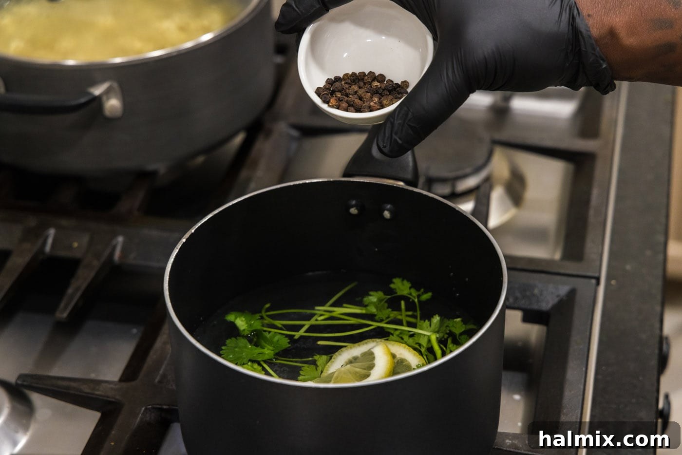 Adding whole black peppercorns to a saucepan already containing cilantro stems, lemon slices, and garlic salt in water