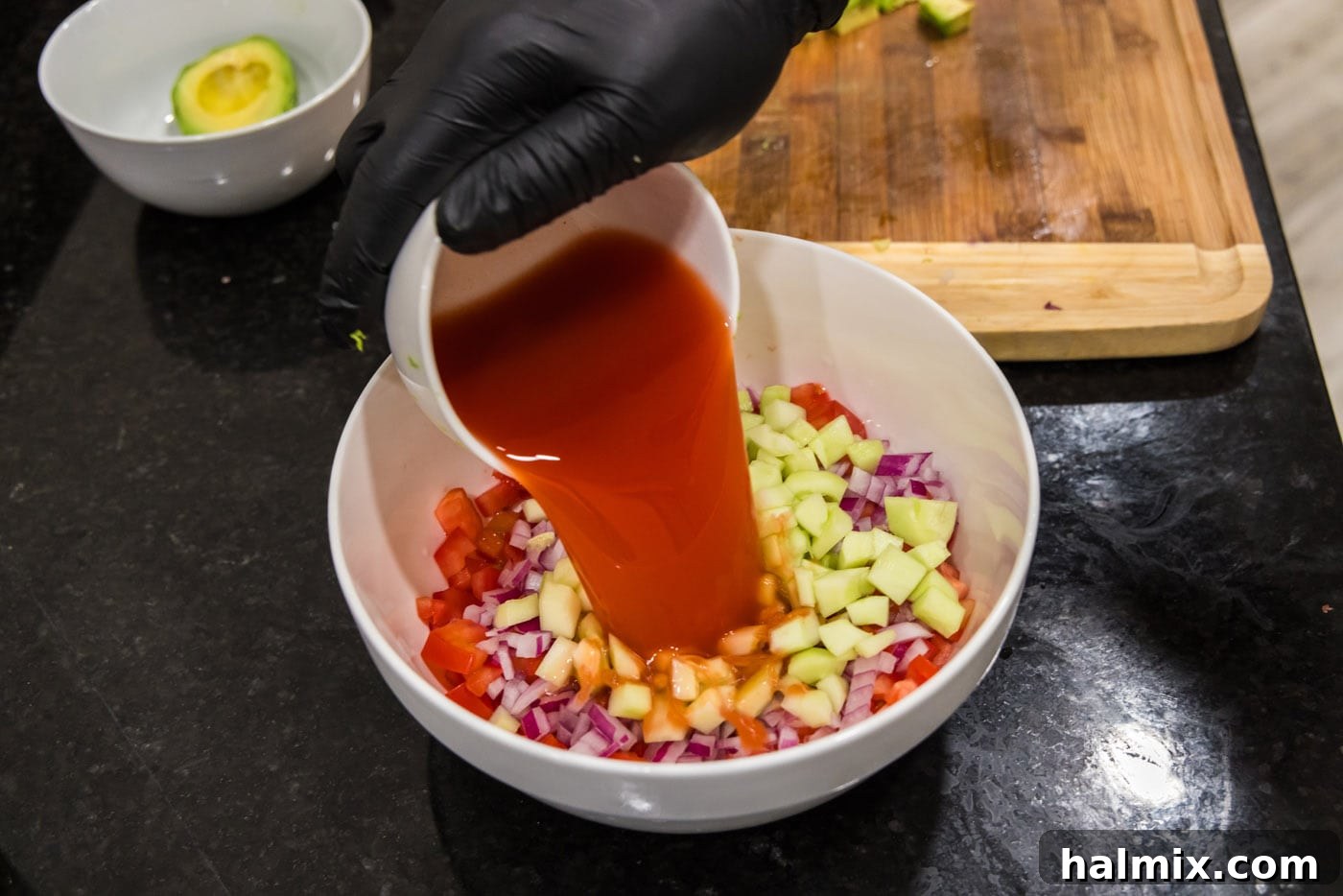 Pouring Clamato juice into a large mixing bowl filled with chopped vegetables, marking the beginning of the broth assembly