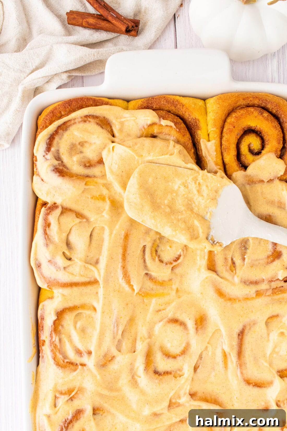 Rich pumpkin cream cheese frosting being generously spread onto warm pumpkin cinnamon rolls in a baking dish