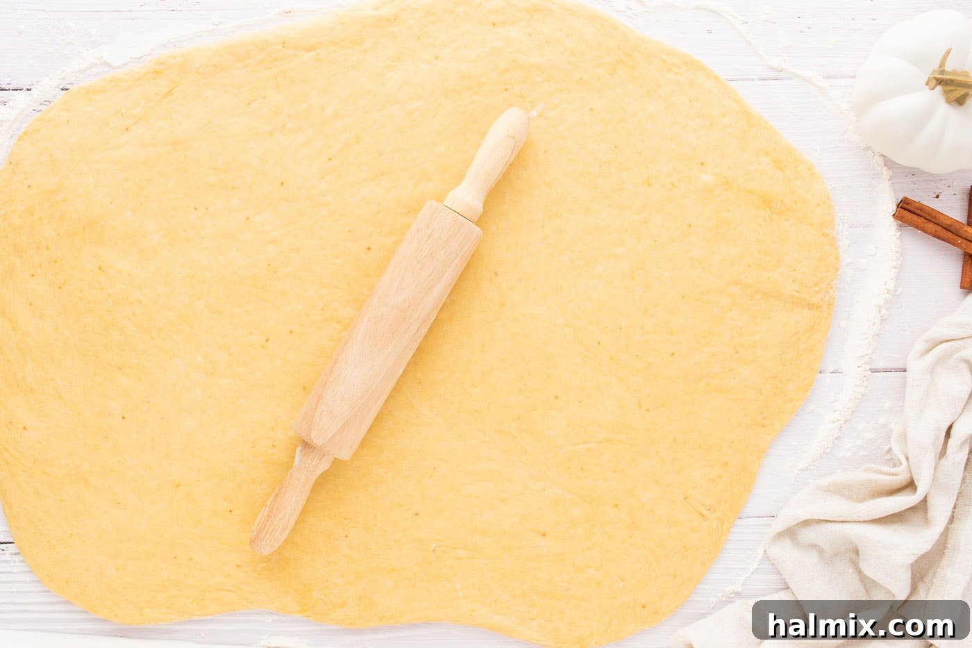 Hands using a rolling pin to flatten cinnamon roll dough into a large rectangle on a floured surface
