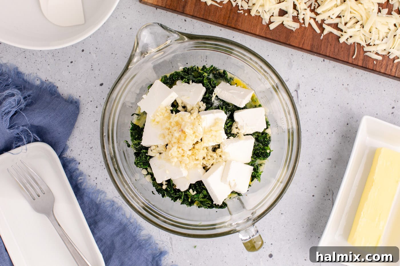Cubed cream cheese, minced garlic, and shredded pepper jack cheese being added to a bowl with eggs and spinach, illustrating the next step in preparation.