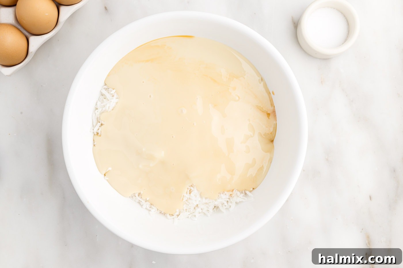 Sweetened condensed milk being poured over shredded coconut and vanilla extract in a bowl, just before mixing.
