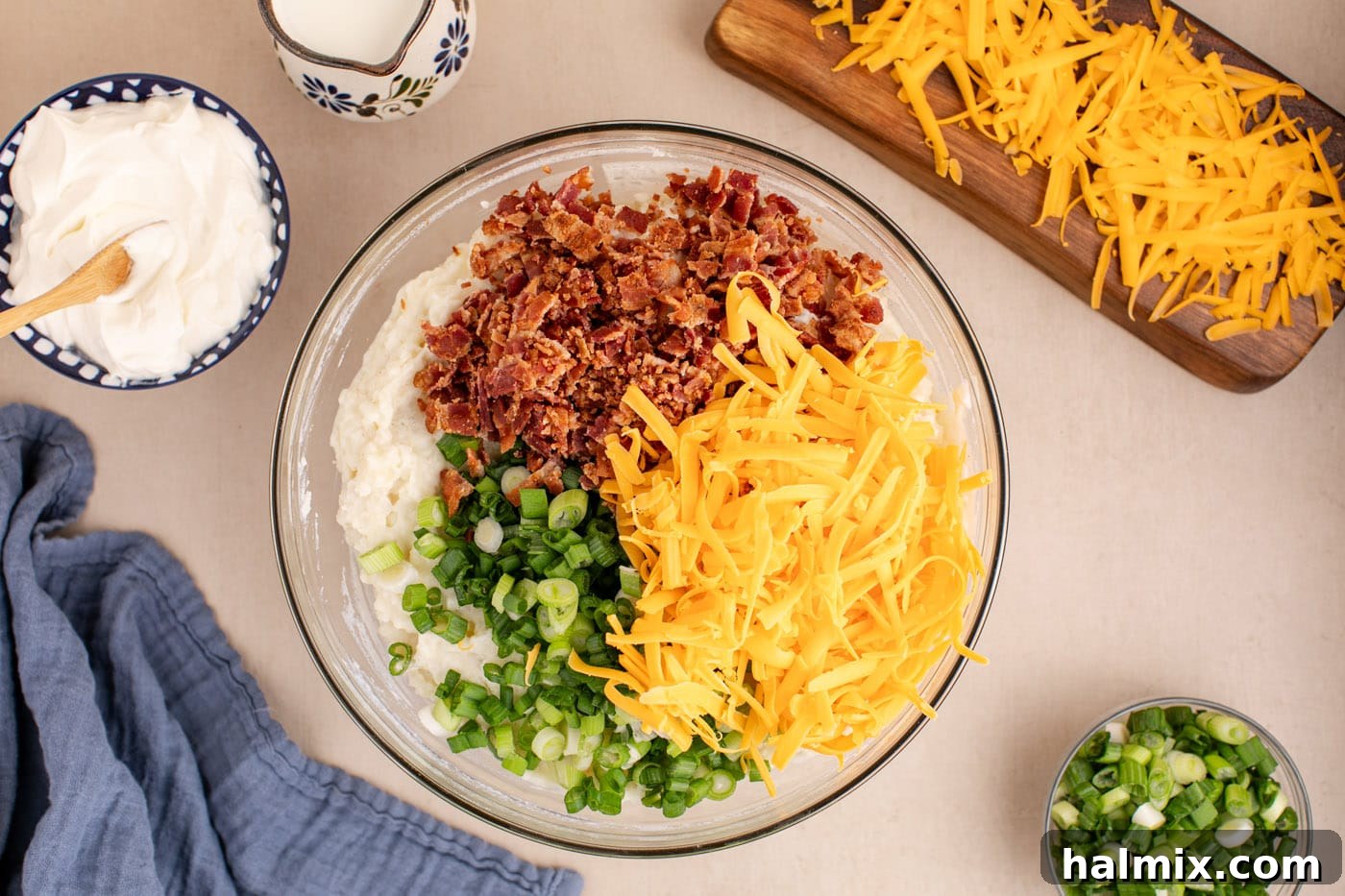 Decadent Twice Baked Potato Casserole 9 Crumbled bacon, shredded cheese, and sliced green onions being added to the creamy potato mixture in a bowl.