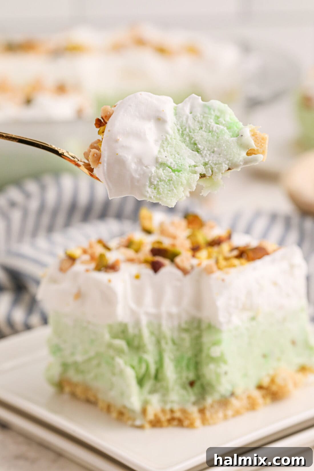 A delectable slice of Pistachio Ice Cream Cake with a fork, showing its distinct layers on a white plate. A larger slice remains on the plate behind it.