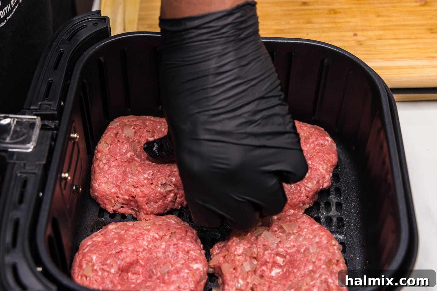 A thumb pressing a shallow indent into the center of a burger patty placed in an air fryer basket, a technique to prevent puffing.