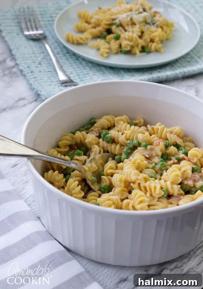 A close-up of a bowl filled with creamy rotini pasta, bright green peas, and pieces of crispy bacon, garnished with fresh herbs.