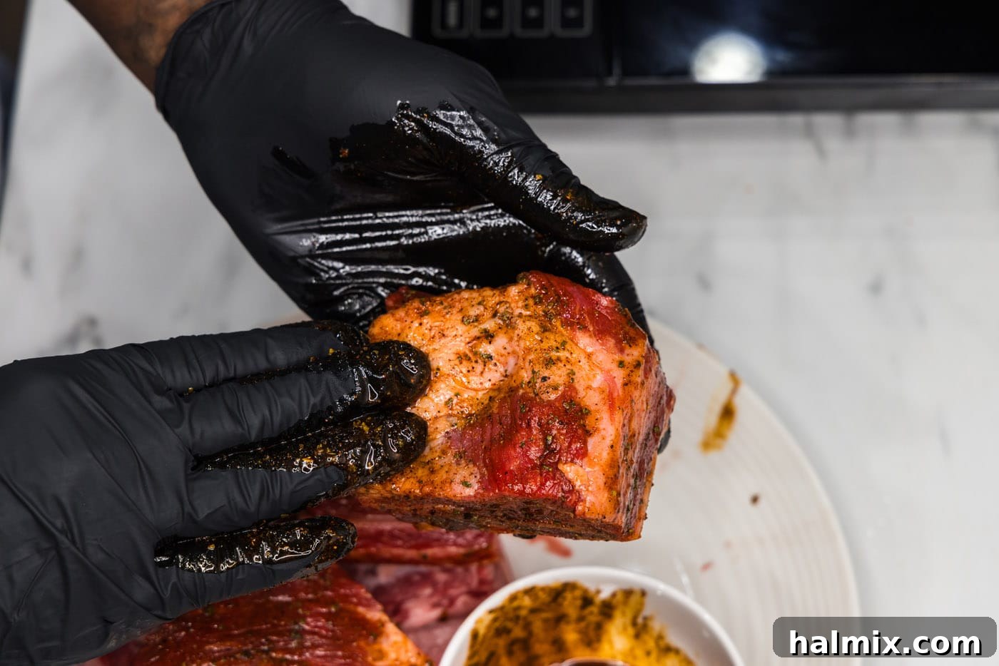 Gloved hands are shown rubbing a mixture of olive oil and seasoning all over raw short ribs, ensuring full coverage.