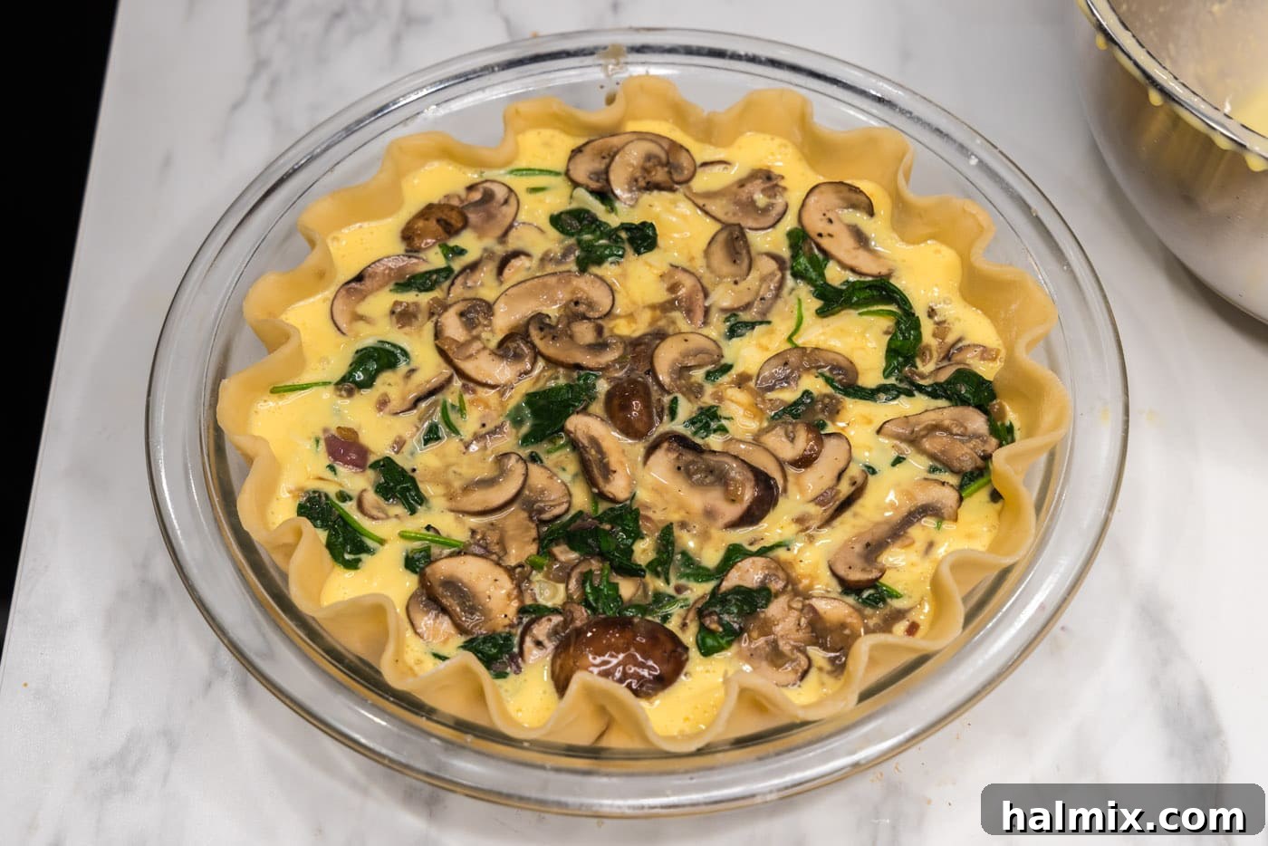 The final layer of spinach and mushrooms sprinkled over the egg filling in the pie plate, completing the assembly before baking.