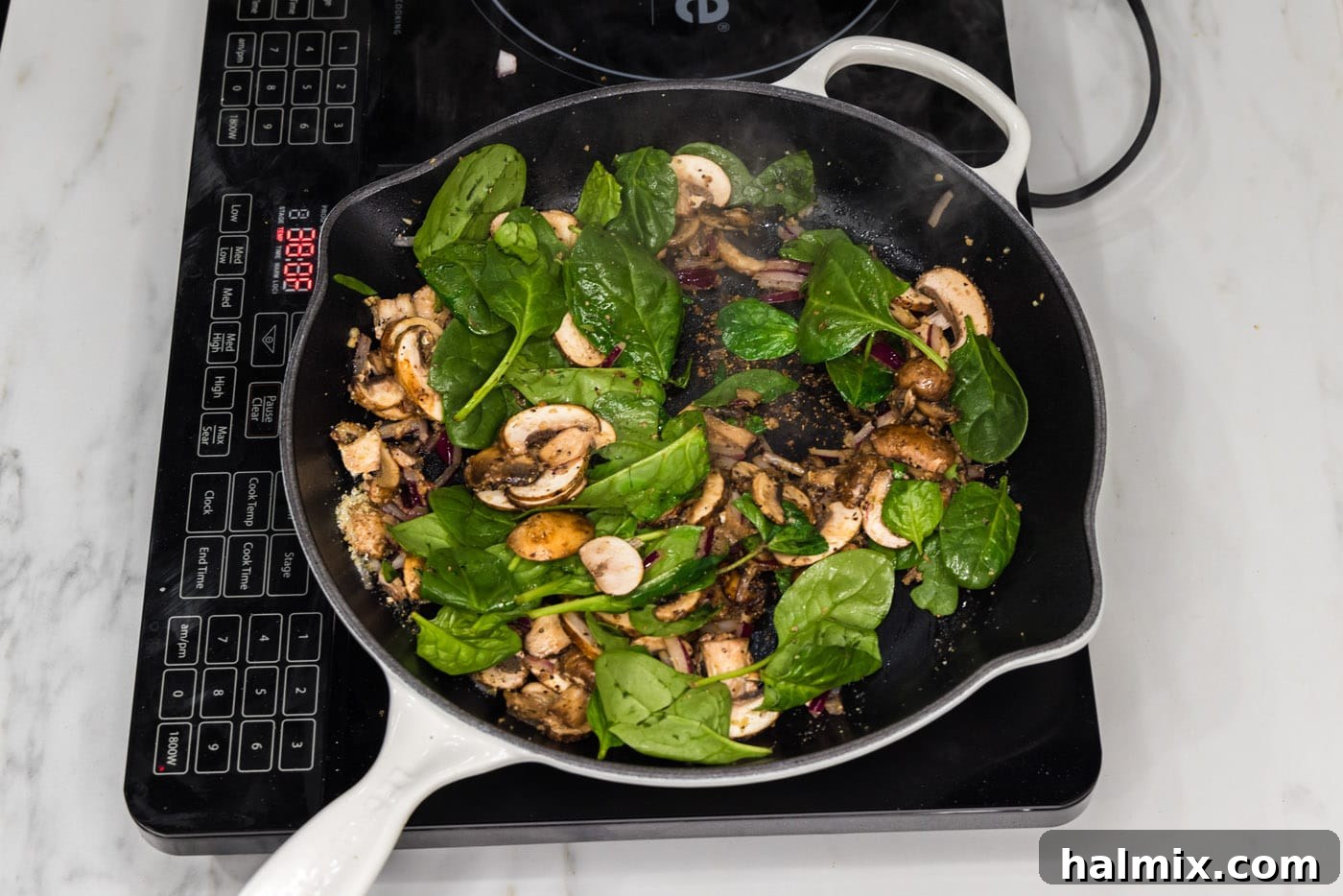 Fresh spinach leaves being added, handful by handful, to the skillet with sautéed mushrooms and onion, beginning to wilt.