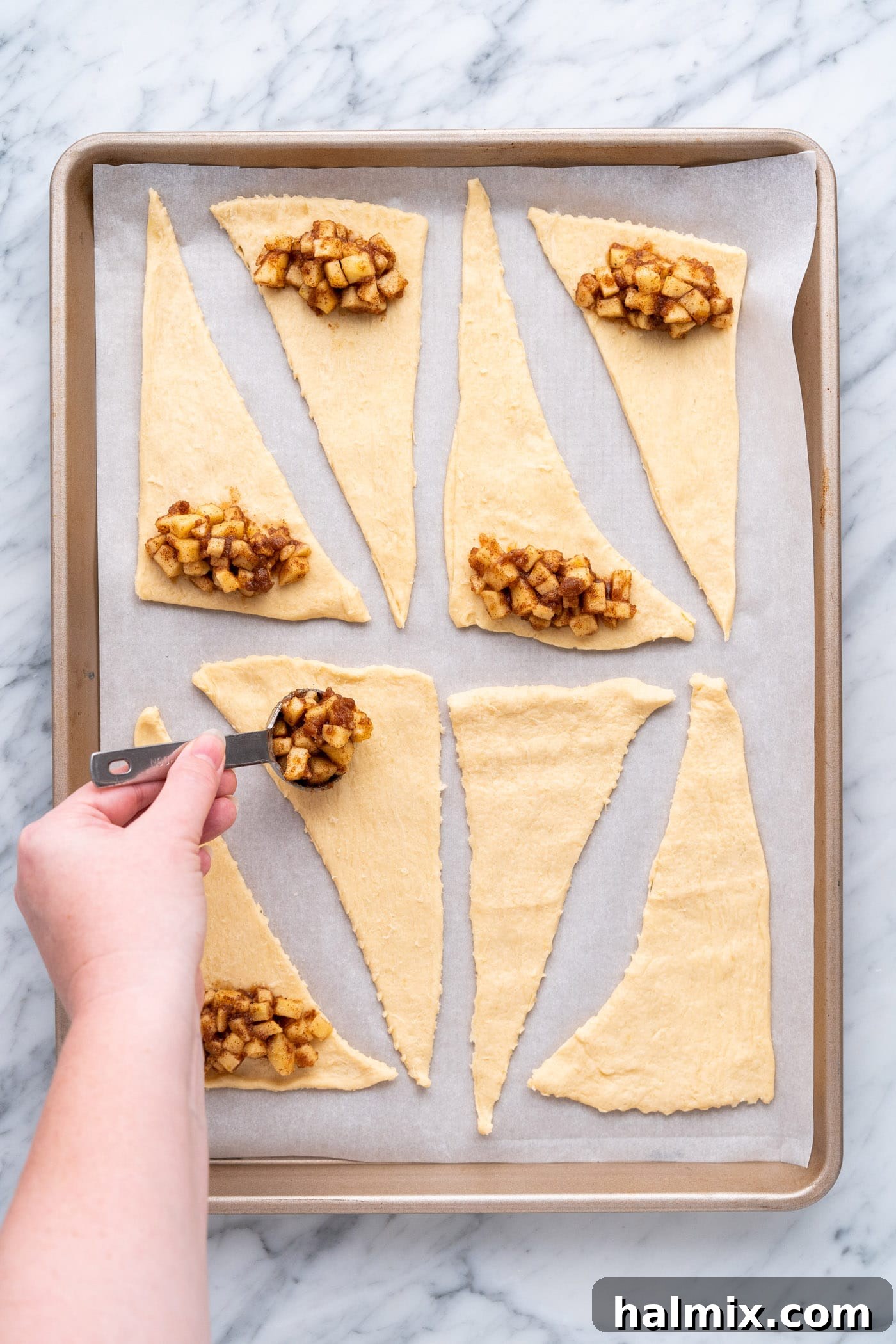 Scooping cinnamon apple mixture onto the wide end of crescent roll triangles on a baking sheet.