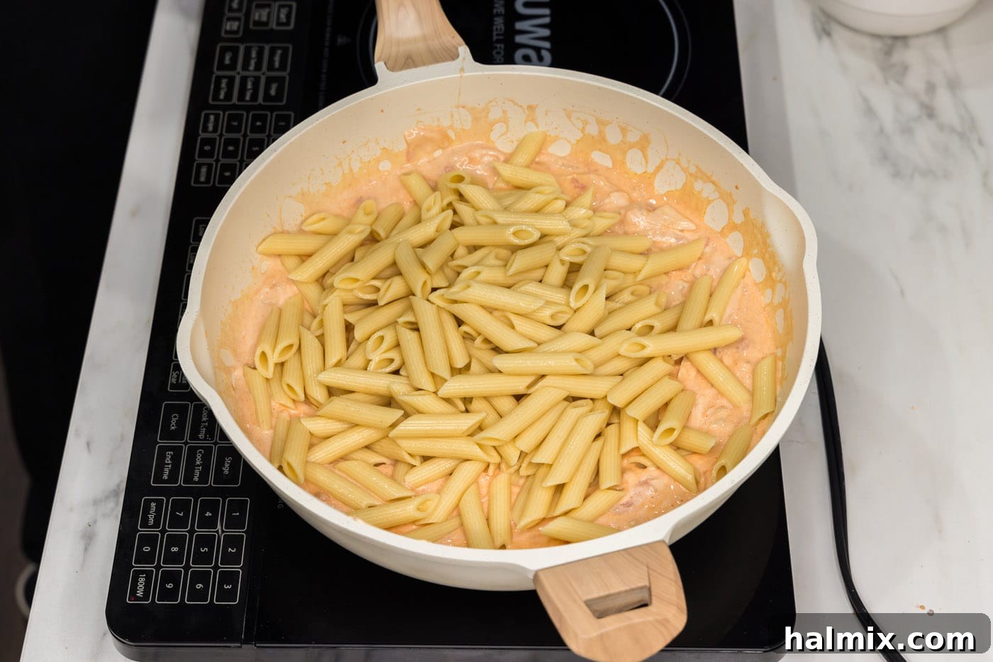 Cooked penne pasta being added to the skillet with the creamy buffalo chicken sauce, ready to be combined.